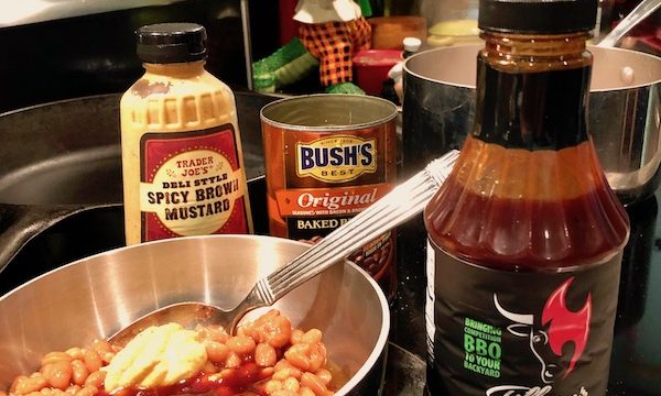 A pot of beans on the stove with mustard and bbq sauce added. Surrounding the pot are a bottle of Tillman's Apple Chipotle BBQ Sauce, an empty can of Bush's Original beans and a bottle of Trader Joe's Spicy Deli Style mustard.