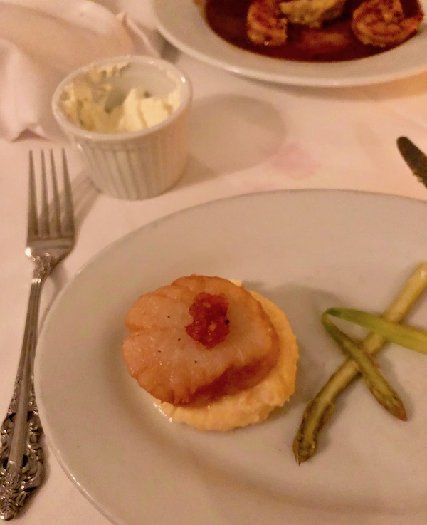 A white plate with grits topped with a sea scallop and shaved asparagus at Brigtsen's in New Orleans Revisited