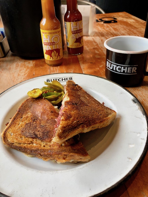 A white plate with a grilled sandwich and pickles on top for Lunch at Cochon Butcher in New Orleans