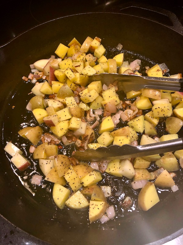 A cast iron skillet with diced potatoes and shallots cooking in oil with a pair of tongs on top for my Andouille Hash with Fried Egg.