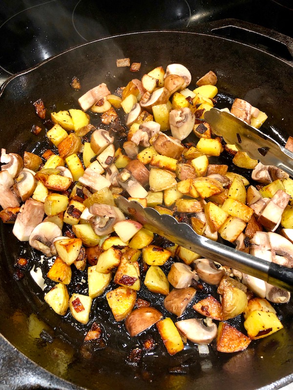 A cast iron skillet with diced mushrooms, potatoes and shallots cooking in oil with a pair of tongs on top for my Andouille Hash with Fried Egg.