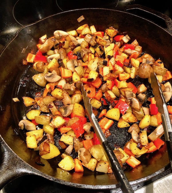 A cast iron skillet with diced, red bell pepper, mushrooms, potatoes and shallots cooking in oil with a pair of tongs on top for my Andouille Hash with Fried Egg.