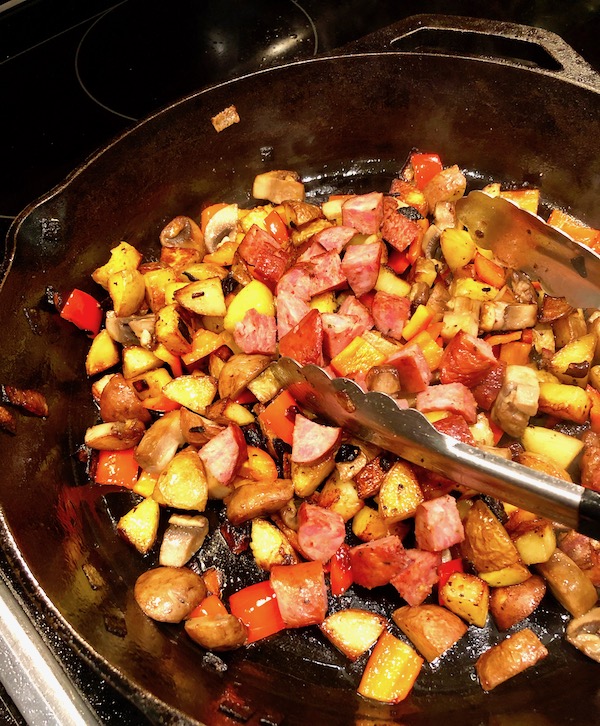 A cast iron skillet with diced, red Andouille sausage, bell pepper, mushrooms, potatoes and shallots cooking in oil with a pair of tongs on top for my Andouille Hash with Fried Egg.