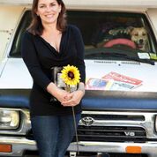 A woman holding a sunflower standing in front of a car with a dog in the drivers seat from Pink Steering Wheel Chronicles