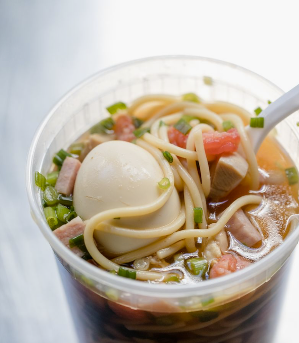 A plastic disposable glass filled with Yakamein Soup. a noodle soup and a hard boiled egg.