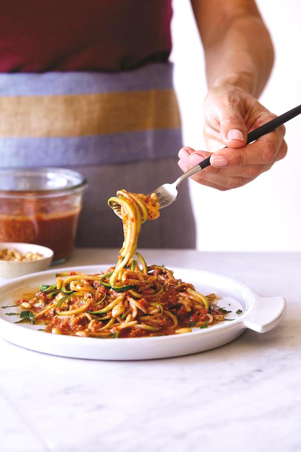 A white pasta bowl holding zucchini noodles with romesco sauce; and a woman hand holding up a forkful of noodles from Eat Like You Give A Fork