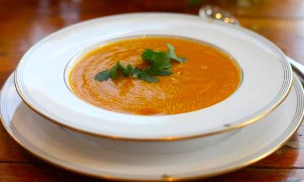 A gold rimmed soup bowl on a matching plate filled with carrot soup from Jewish High Holiday Cooking