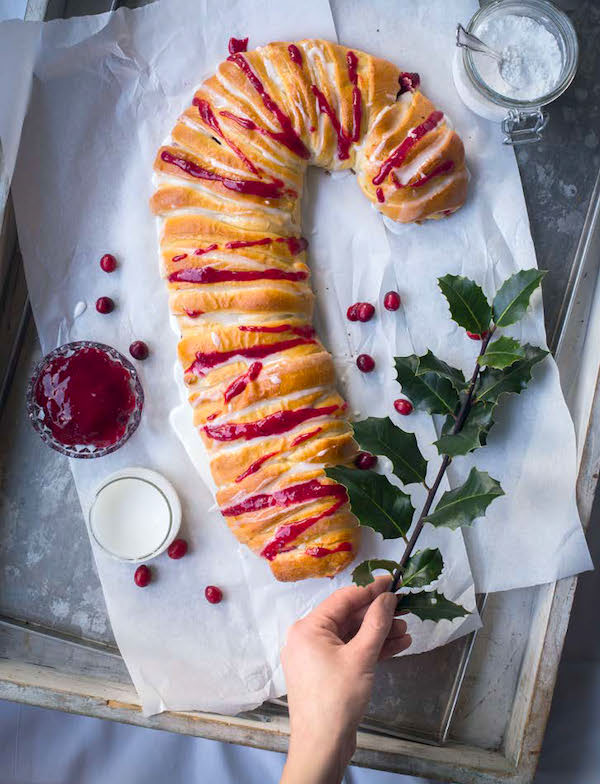 A parchment paper covered baking pan topped with bread in the shape of a candy cane and scattered with cranberries and a branch of holly from New York Christmas Baking