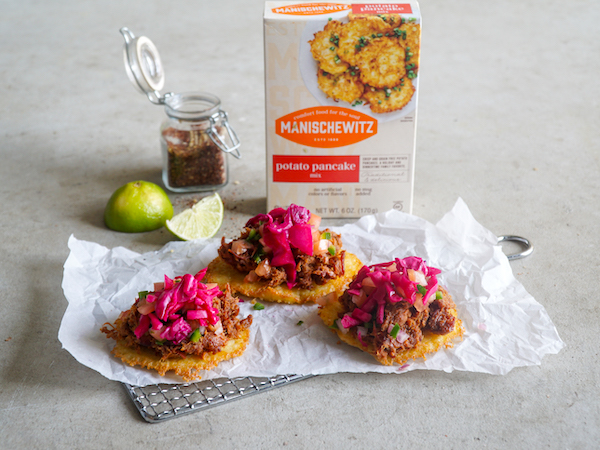 A grey cloth topped with a box of Manischewitz potato pancake mix, limes, potato pancakes topped with pulled brisket and pickled red onion from Pulled Brisket Taco Latke for Hanukkah
