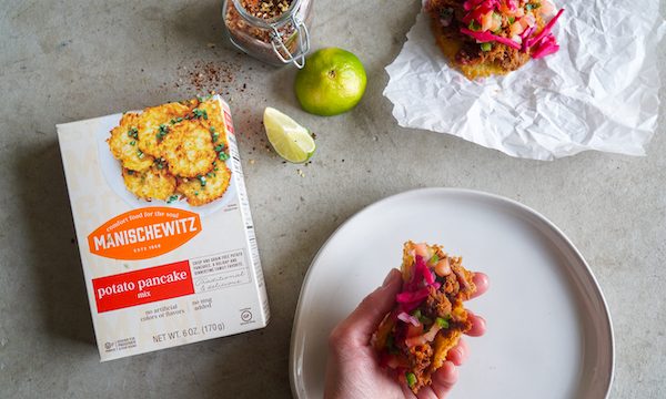 A grey cloth topped with a box of Manischewitz potato pancake mix, limes, potato pancakes topped with pulled brisket and pickled red onion from Pulled Brisket Taco Latke for Hanukkah