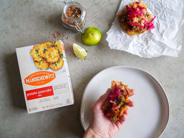 A grey cloth topped with a box of Manischewitz potato pancake mix, limes, potato pancakes topped with pulled brisket and pickled red onion from Pulled Brisket Taco Latke for Hanukkah
