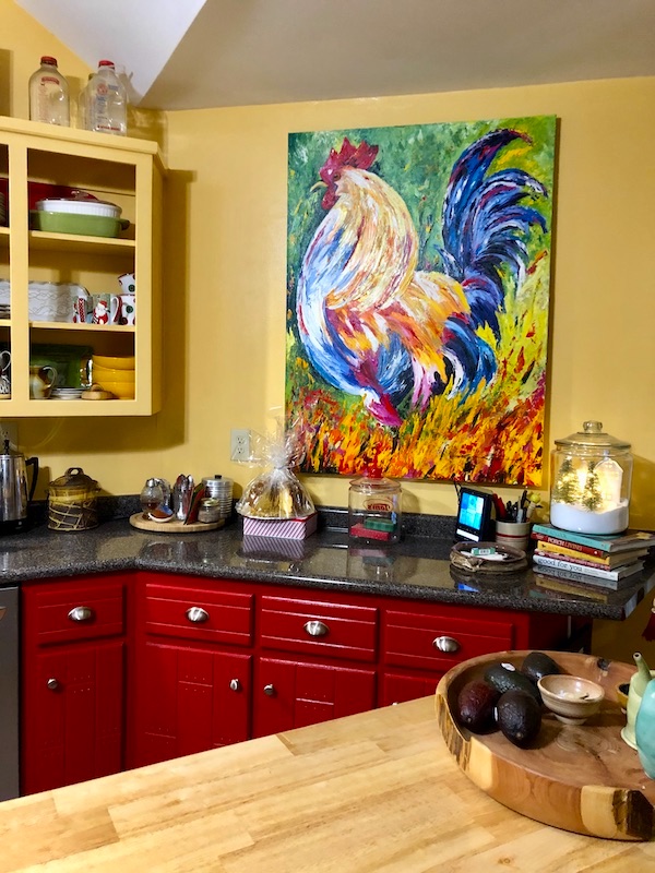 A country style kitchen with red cabinets and a large colorful painting of a rooster