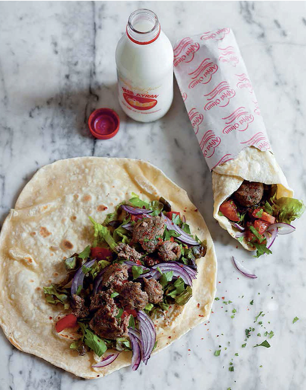 A flatbread filled with meat and veggies on a white marble table along with a rolled sandwich wrapped in paper and a bottle of milk from Istanbul Cult Recipes