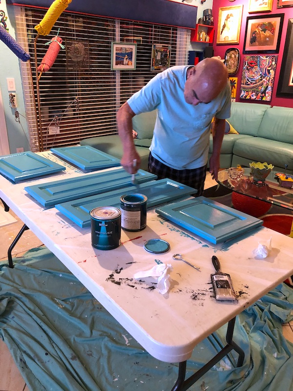 A man painting cabinet doors teal on a table in a living room from Our RV Renovations Update