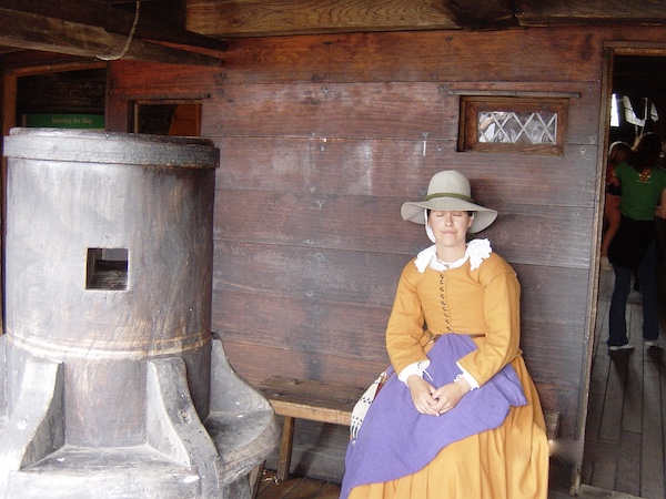 A woman in a gold pilgrim dress with purple apron is resting on a bench at Plymouth Massachusetts