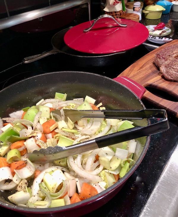 A braising pan on a stove top filled with vegetables and a pair of tongs for Red Wine Braised Beef