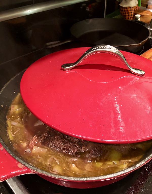 A partially covered red braising pan filled with meat and vegetables for Red Wine Braised Beef