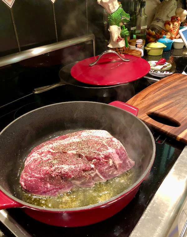 A shallow red pan on a stove top with meat searing in olive oil for Red Wine Braised Beef