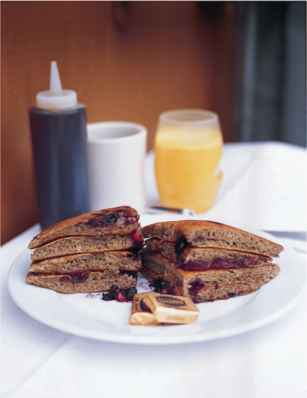 A plate of stacked pancakes with squeeze bottle of syrup, a white mug and glass of OJ from Los Angeles Cult Recipes