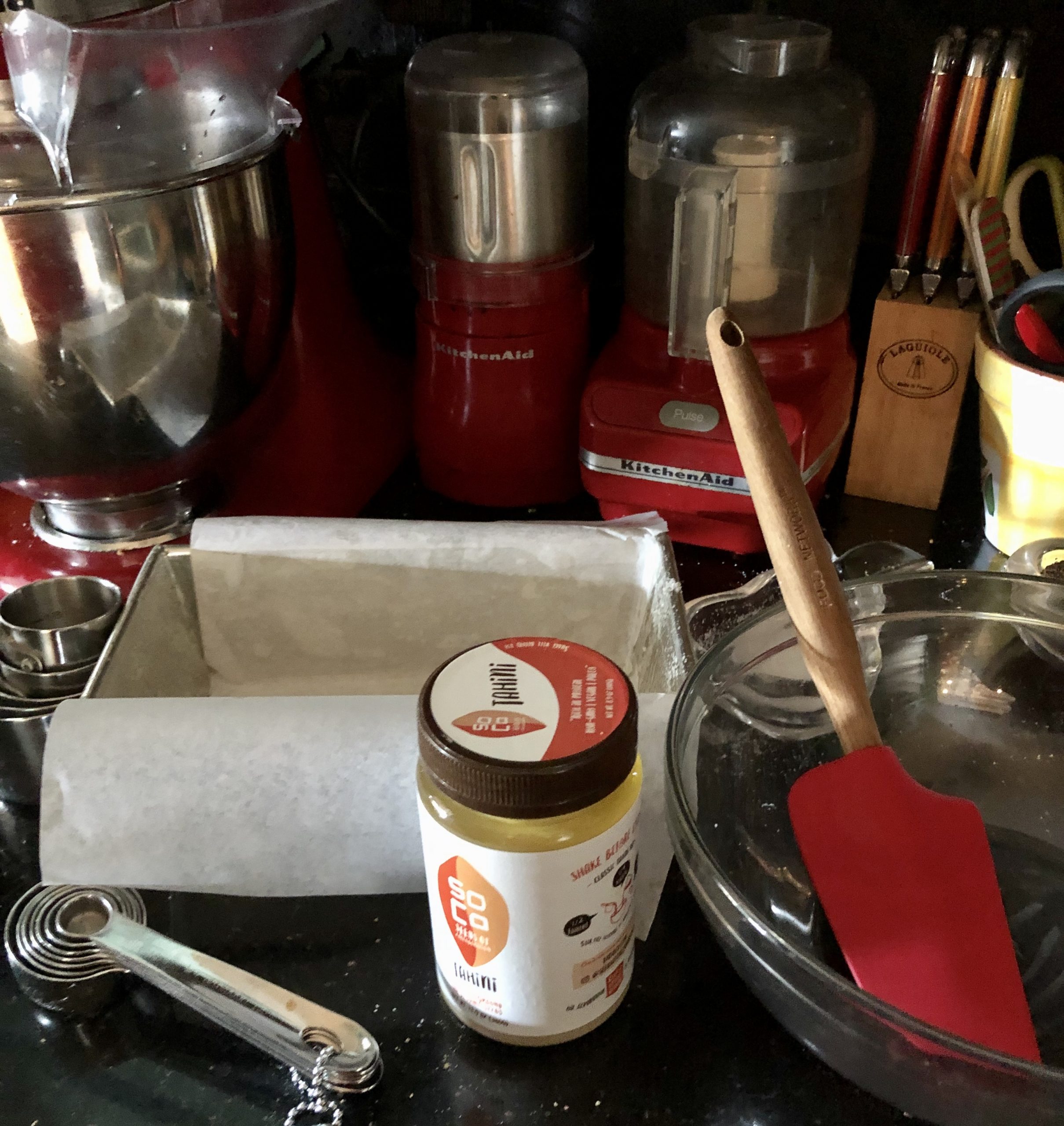 A parchment lined loaf pan, jar of tahini, glass bowl and red silicone spatula for my Salted Chocolate Covered Halvah