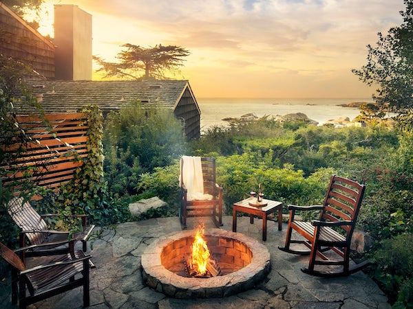 A fire pit surrounded by chairs with a view of the ocean, trees and an Inn from Hyatt Carmel Highlands Reopened.