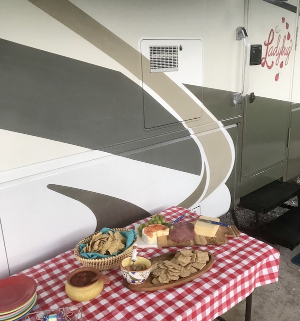 An RV with a table set by its side cover in red and white checked cloth and platters of cheese and crackers from Living Our Dream