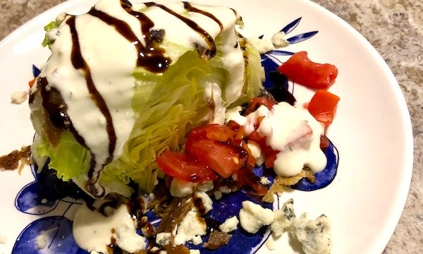 A white and blue plate topped with an iceberg lettuce wedge, bacon, tomato, blue cheese crumbles and balsamic drizzle from RVing With Family