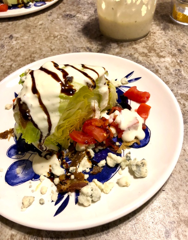 A white and blue plate topped with an iceberg lettuce wedge, bacon, tomato, blue cheese crumbles and balsamic drizzle from RVing With Family
