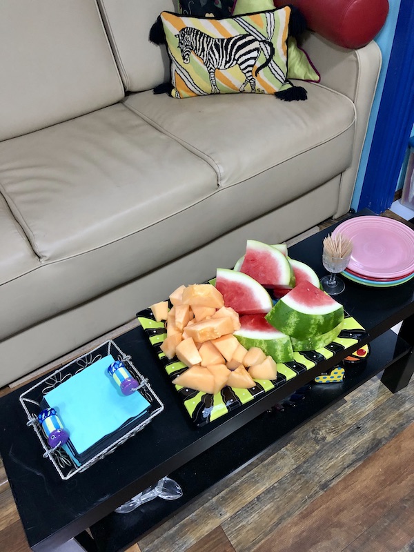 A platter of cantaloupe and watermelon, aqua napkins and small plates on a black table in front of a beige sofa at North River Campground NC