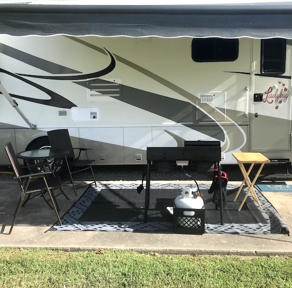 The side of a white and tan RV with awning out, patio rug, grill, chairs and table at North River Campground NC