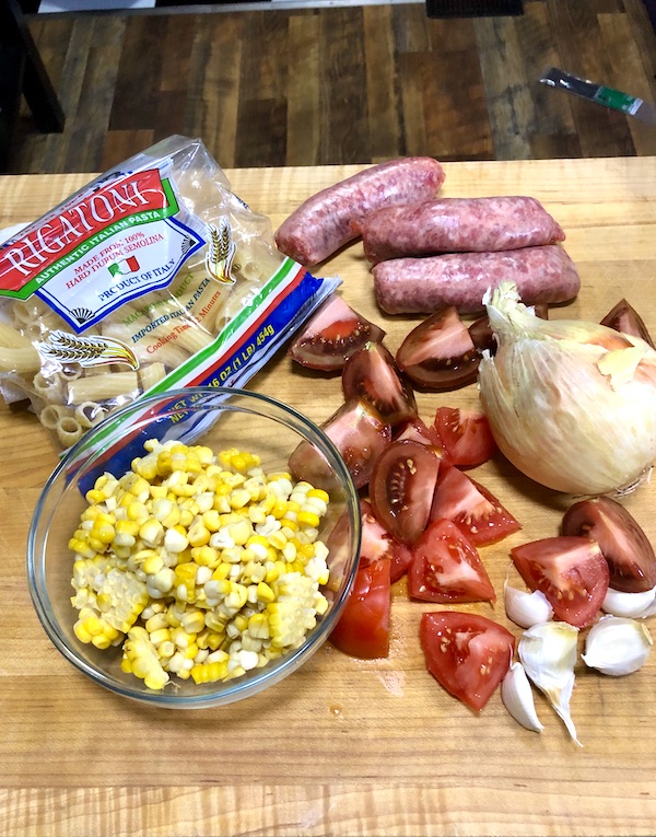 A wood board topped with a bag of pasta, a bowl of corn, wedges of tomatoes, garlic cloves, an onion and sausages for my Rigatoni with Green Cut Sausage