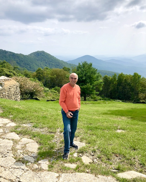 A man in an orange sweater standing on a lawn with mountains behind him near Boone North Carolina