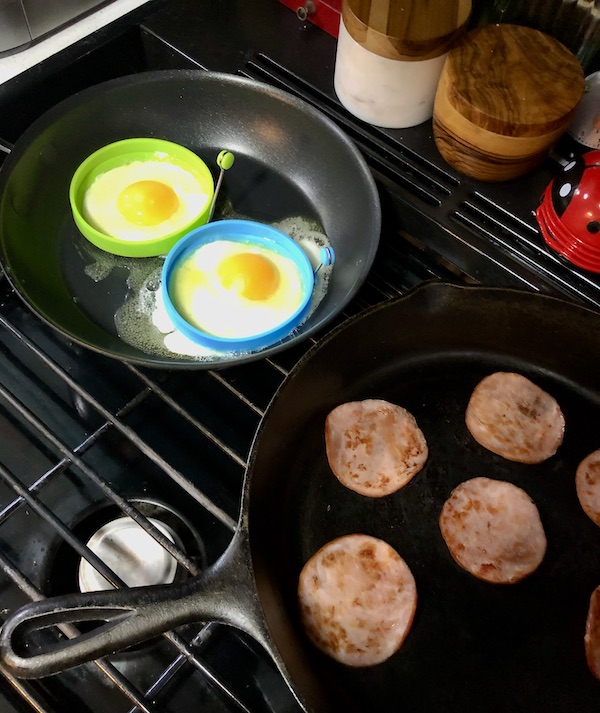 A gas stove top with two sauté pans. One pan with eggs and the other with round bacon for Breakfast Sandwiches