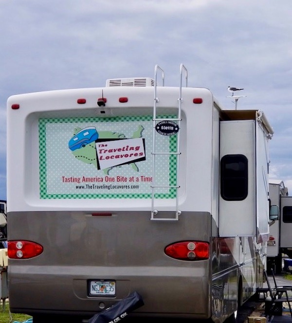 A white and tan Class A RV with The Traveling Locavores logo on the back and a seagull perched on the antenna at Delaware Seashore State Park