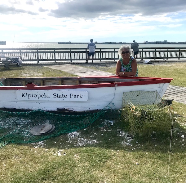 A woman behind a white row boat with fishing pier behind her at Kiptopeke State Park in Virginia