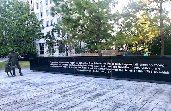 A black wall with white lettering depicting an oath of government office in Jackson Mississippi. A sculpture of a father and daughter stands in front.