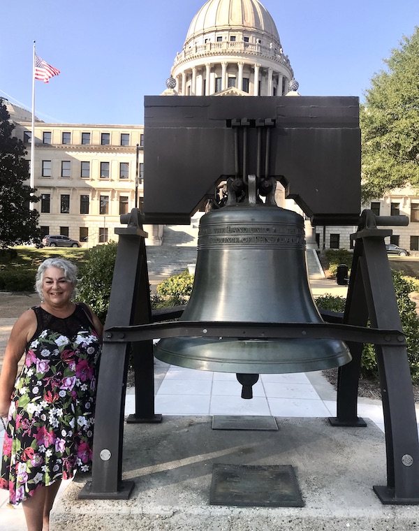 A woman in a print dress standing next to a replica of the Liberty Bell in from of the Capitol Building in Jackson Mississippi