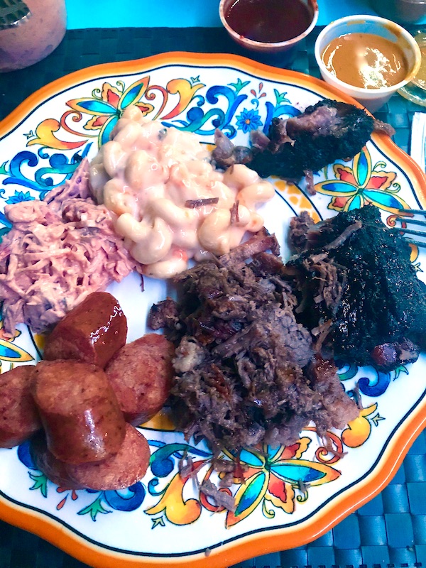 A colorful plate topped with mac and cheese, ribs, shredded meat, sliced sausage and pink coleslaw from a BBQ restaurant near the Mississippi Civil Rights Museum