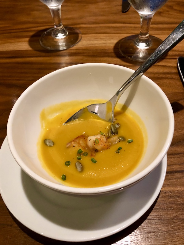 A beige soup bowl filled with orange soup and garnished with shrimp and greens with a soup spoon in the bowl at Sue Zemanick's Zasu