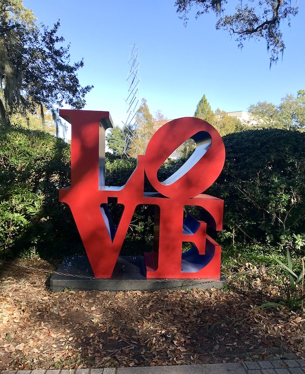 A larg red LOVE sculpture nestled in the bushes at the Bestoff Sculpture Garden