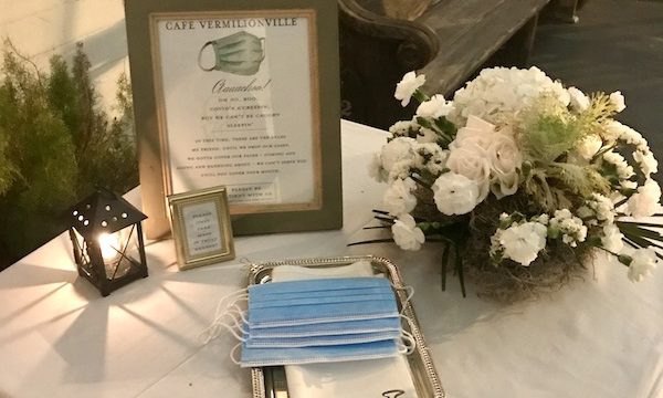 A table with white tablecloth, white flowers, disposable masks and a framed notice to take a mask if you don't have your own sits on the front porch of Café Vermillionville Lafayette Louisiana