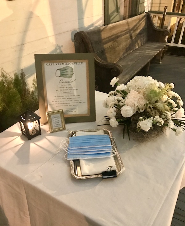 A table with white tablecloth, white flowers, disposable masks and a framed notice to take a mask if you don't have your own sits on the front porch of Café Vermillionville Lafayette Louisiana