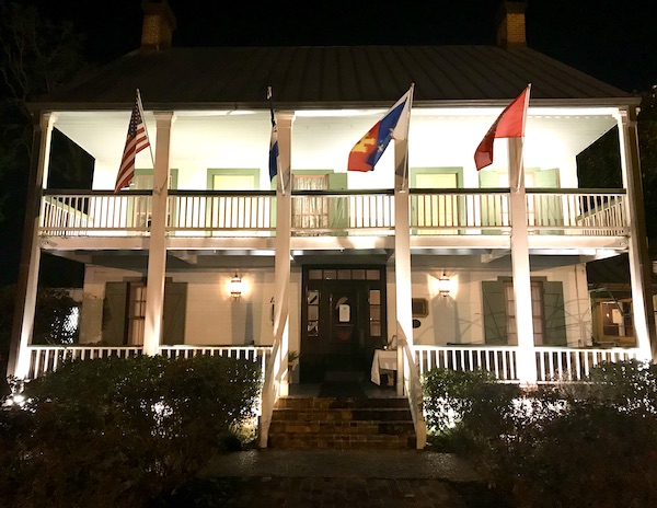 The front of a two story restaurant with a front porch, columns and flags is Café Vermillionville Lafayette Louisiana