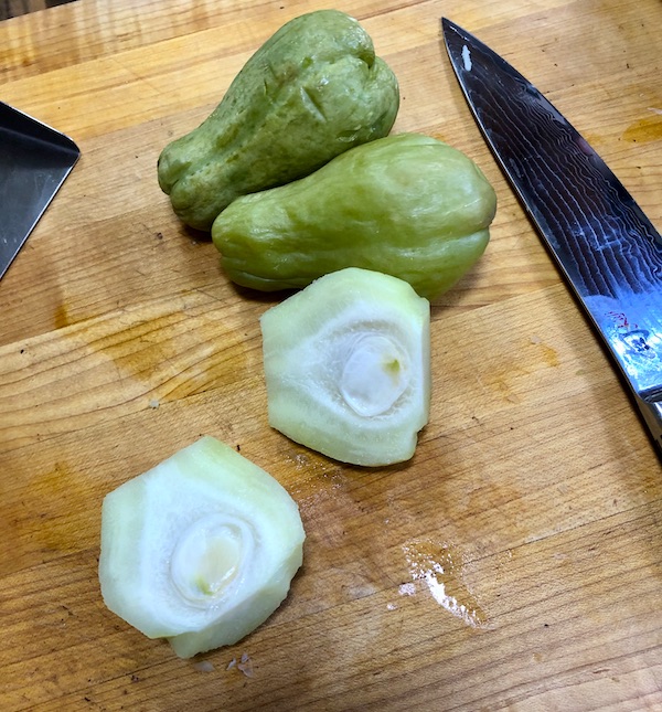 Seasonal Ingredient Mirliton sitting on a cutting board two unpeeled and one peeled and cut in half.