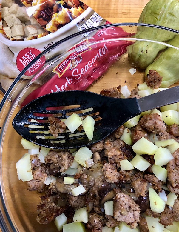 Seasonal Ingredient Mirliton diced in a bowl with sausage and mushrooms, with a black slotted spoon resting on top.