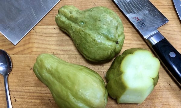 Seasonal Ingredient Mirliton are shown as three green pear shaped vegetables, one partially peeled revealing white interior. The vegetables are sitting on a wood cutting board along with a small spoon and chef's knife.