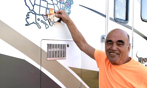 A man in an orange tee shirt placing a Texas at Last! sticker on a map on the side of an RV