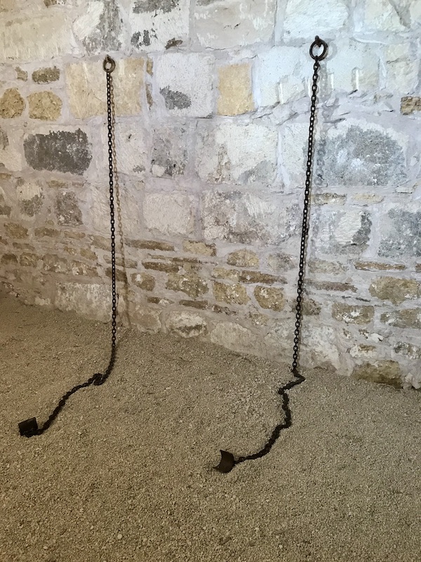 A stone wall with chains and shackles hanging and laying on the dirt floor at Fort Stockton Texas