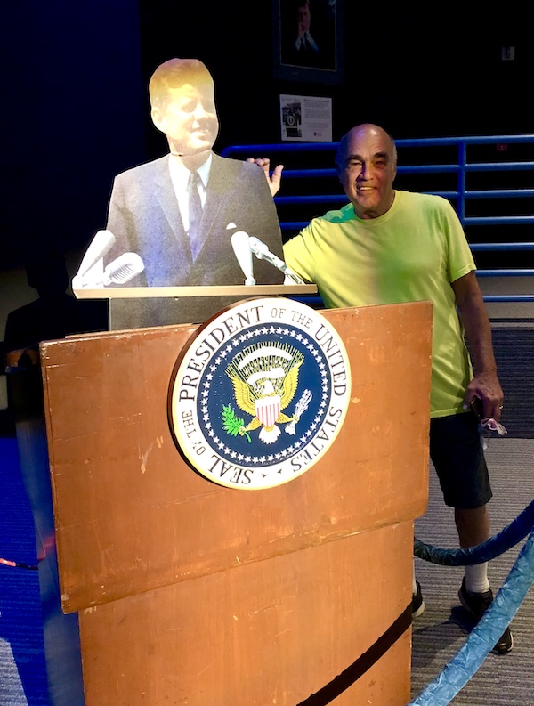A man in a yellow tee shirt at a podium next to a replica of JFK at NASA Space Center Houston
