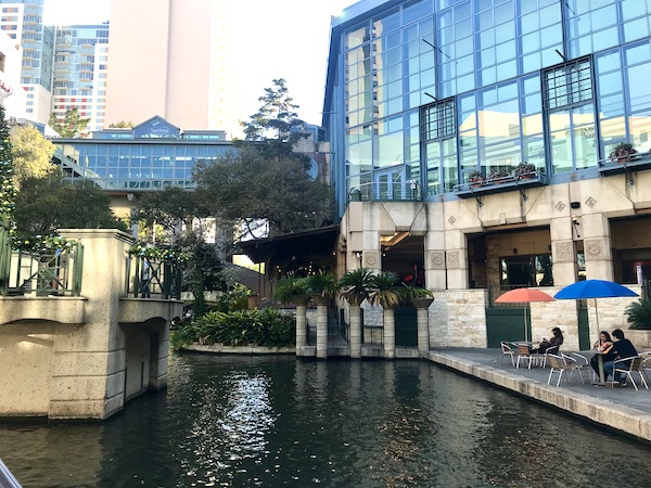 River, stores and people at umbrella tables at the beginning of our San Antonio River Cruise